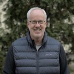 The smiling face of a white man with grey hair, a receding hairline and glasses.